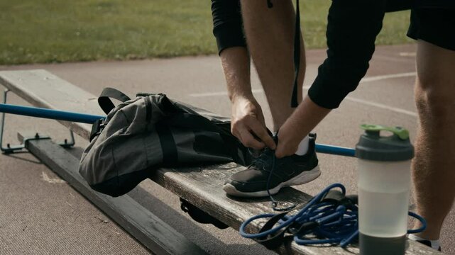 Medium closeup of hands and feet of unrecognizable male athlete with congenital disability, missing hand, swiftly tying up laces on trainers before workout at outdoor stadium