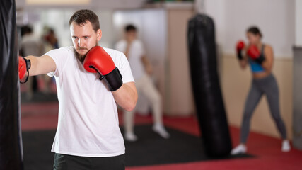 Man practices blows in gloves on a punching bag in the boxing section. European came to a group boxing training and learns punches