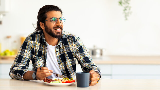 Happy middle-eastern young bearded man in casual outfit and stylish glasses enjoying healthy lunch at home, sitting at kitchen table, eating breakfast, looking at copy space, healthy deit concept