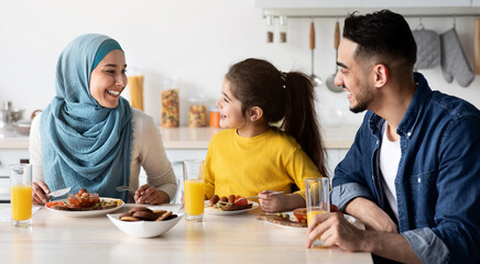 Domestic Breakfast. Happy Middle Eastern Family Of Three Eating Together In Kitchen, Cheerful Young Arab Parents And Little Daughter Sitting At Table, Enjoying Tasty Food, Chatting And Laughing