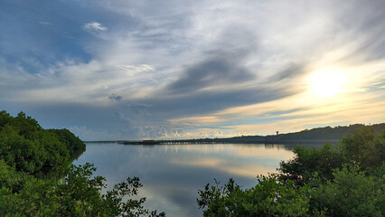 A serene view of mangroves reflecting on calm water under a dramatic sky at sunrise or sunset. The lush greenery contrasts with the soft hues of the sky. Barranquilla