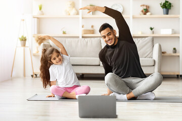 Smiling arab father and little daughter having online yoga class, exercising at home together,...
