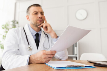 Medicine and Prescription Concept. Pensive male general practitioner holding paper, reading medical report, sitting at desk in his office at clinic. Doctor thinking about test results and treatment