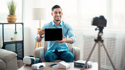 Arab guy influencer shooting video review of modern tablet computer on professional dslr camera at home, mockup for design. Cheerful blogger advertising innovative tech product, streaming live on web