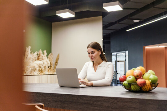Professional woman working on laptop in modern office with fresh fruit display nearby during daylight