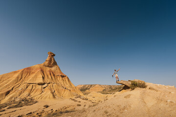 A traveler stands on the edge of a rocky cliff, arms stretched wide, overlooking the breathtaking desert formations of Bardenas Reales under a vibrant blue sky.
