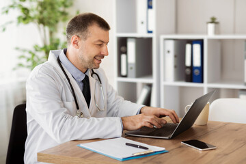 Fototapeta premium Healthcare And Telemedicine Concept. Portrait of smiling middle aged general practitioner using computer laptop, typing on keyboard, writing report, sitting at desk in his office at clinic