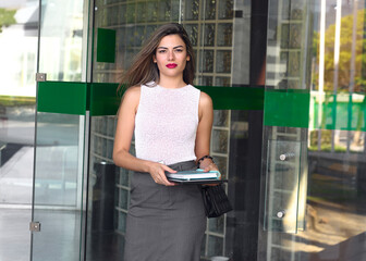 Professional woman exits building with a tablet on a sunny day in an urban setting, showcasing...