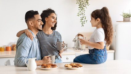 Happy arab family enjoying breakfast at cozy kitchen, little girl daughter sitting on table, drinking milk and having conversation with hugging smiling father and mother drinking morning coffee