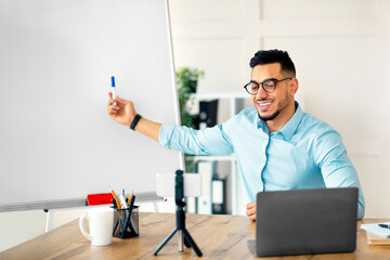 Cyberlearning, remote education concept. Happy Arab guy teacher giving online English lesson, using smartphone and laptop, pointing at blackboard with grammar rules at home office