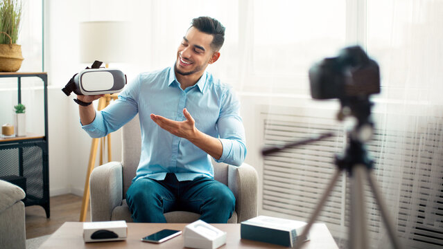 Tech product review. Arab guy vlogger presenting VR headset, recording video about innovative device for technology blog, using camera at home. Trendy influencer filming brand advertising promo