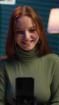 Vertical video Young content creator records introductory video with smartphone, waving hand, inviting viewers to subscribe to channel. Media star uses mobile phone to film herself connecting with