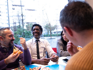 Seen from the perspective of a fourth person in a beige sweater, three colleagues enjoy pizza and laugh together, with a phone on the table. The woman appears slightly more serious