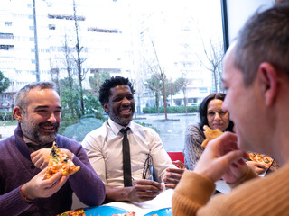 From the perspective of a man in a beige sweater, three colleagues with pizzas look at him while laughing. A phone is on the table, and rain falls outside the bright office window