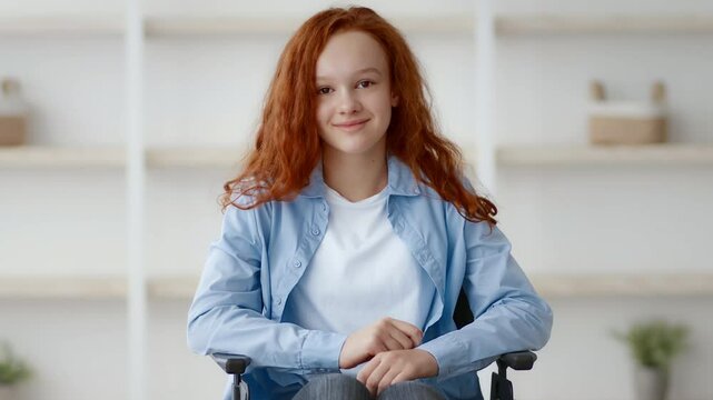 A teenage girl with red hair sits in her wheelchair at home, smiling brightly. The background features simple decor, highlighting her cheerful demeanor and comfort in this space.