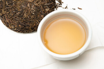 Chawan porcelain bowl with fresh brewed sheng pu-erh tea. Pressed raw pu-erh tea cake on table full of aged tea leaves. Real masterpiece craftsmanship tradition, tea preparation and asian culture