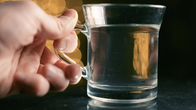 Glass of water placed on bedside table at night. Clear drinking cup with golden bokeh lights background. Evening bedroom atmosphere in macro shot