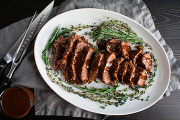Sliced Tri-Tip Roast Beef with Red Wine Glaze: Slices of roast beef with sauce surrounded with sprigs of thyme and rosemary on a ceramic plate