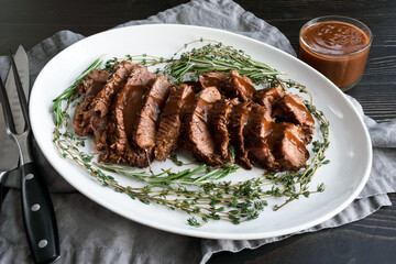 Sliced Tri-Tip Roast Beef with Red Wine Glaze: Slices of roast beef with sauce surrounded with sprigs of thyme and rosemary on a ceramic plate
