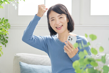 スマホを持つ中年女性　ガッツポーズ　リビング