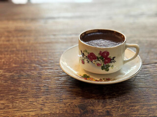 A close-up of a cup of dark coffee in a vintage-style porcelain cup and saucer, resting on a rustic wooden table with a rich, warm brown color.