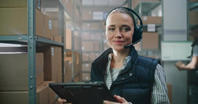 Portrait of Female Logistics Manager Wearing Headset Using Tablet Computer, Looking at Camera in Warehouse Facility with Online Orders. Sorting Center Employee Working in the Background. Close Up.