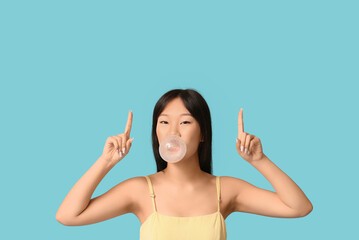 Pretty young Asian woman blowing bubble gum and pointing at something on blue background