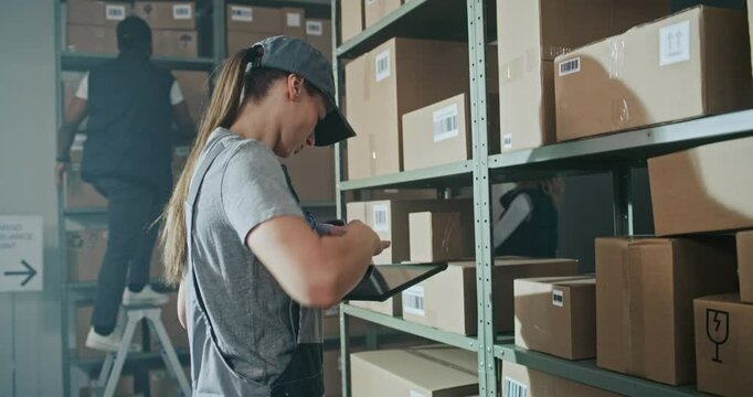Female Warehouse Worker Scans Cardboard Box with E-Commerce Online Goods on Shelf with Scanner, Using Tablet Computer, Takes Parcel for Shipping to Client. Sorting Center of Delivery Service. Handheld