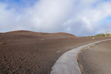 Volcanic desert landscape with a paved trail along black sandy hills with few bushes and plants and blue sky above. Devastation trail, Hawaii volcano national park, Big Island, Hawaii, the USA.