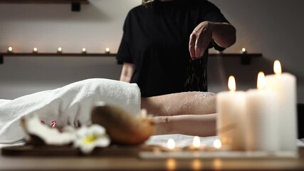 A massage therapist sprinkles coconut flakes on a woman’s back in a serene spa, with a coconut and candles in the foreground, creating a peaceful atmosphere. Perfect for relaxation and rejuvenation.