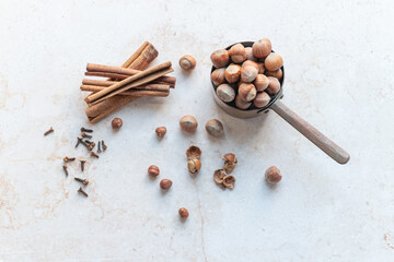 Rustic composition featuring whole hazelnuts in a vintage metal scoop, cinnamon sticks, and cloves scattered on a light marble surface. Ideal for culinary projects, recipes, or product branding.