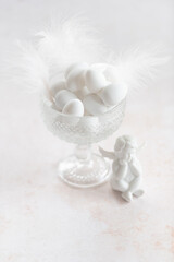 An elegant arrangement of white sugared almonds is in a glass bowl with a delicate pattern, surrounded by soft white feathers. A charming angel figurine adds a sentimental and warm touch.