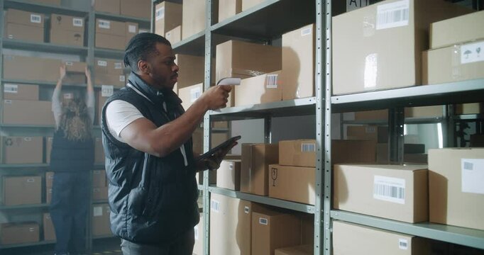 African American Inventory Specialist Walks in Warehouse Retail Facility, Scans Parcels with E-Commerce Online Orders, Checks Delivery Information on Tablet Computer. Sorting Center of Postal Service.