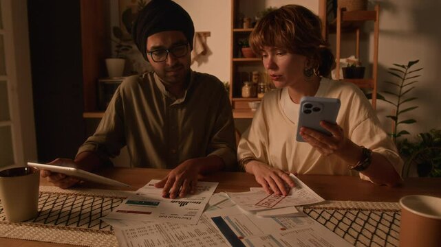 Tilt shot of unpaid utility bills and eviction notice lying on table, diverse husband and wife discussing family budget and solving financial problems, using smartphone and tablet