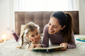 Fototapeta premium Adorable daughter and mother lying in bed and reading a story form a book at home.