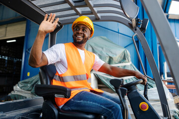 Friendly black forklift driver driving forklift and waving to his colleagues at industrial zone.
