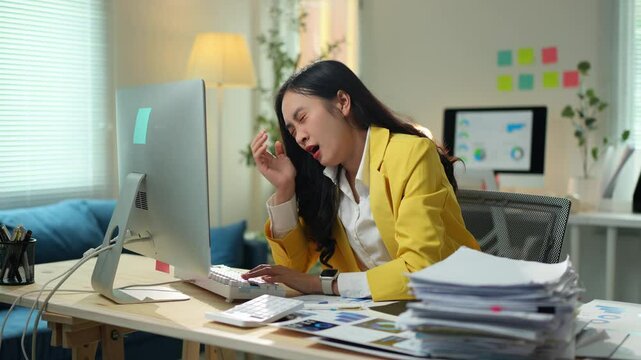 Young asian businesswoman in yellow suit working on computer in office, getting increasingly tired and eventually falling asleep on a pile of paperwork