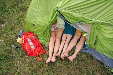 Three pairs of feet are protruding from a green tent, with backpacks resting nearby on a bright,...