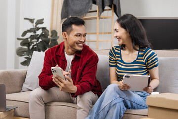 Young couple sitting on the sofa in their new house, happily using a laptop, enjoying each other's company, and creating joyful memories in their fresh, cozy home environment