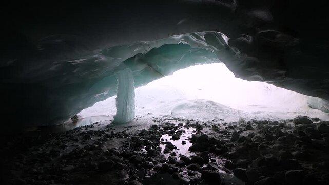 A winter adventurer with a camera appears from the darkness of a glacier cave in the Swiss Alps before inspecting the ice of a giant frozen icicle and taking photos