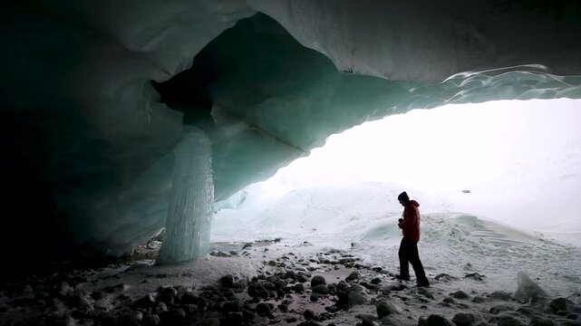 A winter adventurer with a camera appears within a glacier cave in the Swiss Alps to inspect the ice of a giant frozen icicle and take photos