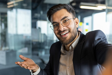 Video call, businessman smiling looking into smartphone camera. Man at workplace inside office in business suit working, using smartphone app for remote conversation.