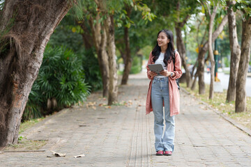 Young asian student or freelancer is walking in a tree-lined street, holding a digital tablet and wearing casual clothing and backpack, enjoying the fresh air and greenery