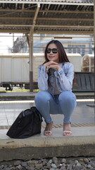 Fit woman in jeans and denim jacket, confidently squatting on a platform near her travel bag