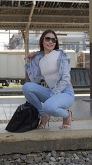 Beautiful woman in denim and high heels, confidently squatting on a platform near her travel bag