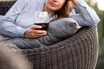 A glass of red alcoholic drink or wine in a woman's hand. A woman is resting on the veranda of a country house. The concept of relaxation, party or celebration.