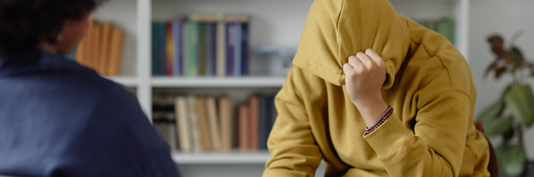 Header image of teenage boy sharing mental health troubles with therapist while sitting on chair and hiding face during one on one session copy space