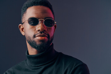 Obraz premium Stylish African man wearing sunglasses and a turtleneck, showcasing confidence against a dark gradient background, perfect for fashion and lifestyle concepts