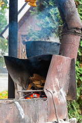 photo shows a boiler on a campfire. Wood is burning in a metal stove from which smoke is rising. Trees and a wooden house can be seen in the background.