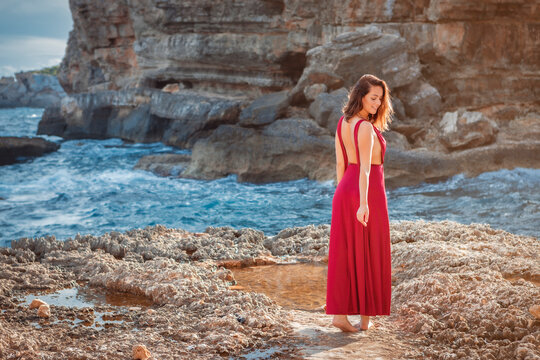 portrait of a young brunette woman standing next to the ocean shore, wearing a bordeaux red dress